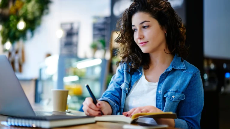 Young woman studying at a café, writing notes in a notebook while working on a laptop with a coffee cup beside her